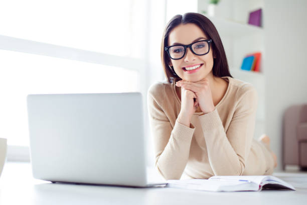 Portrait of cute nice-looking smiling businesswoman in glasses sitting at the table with laptop, notepad on it and holding up her head with hands